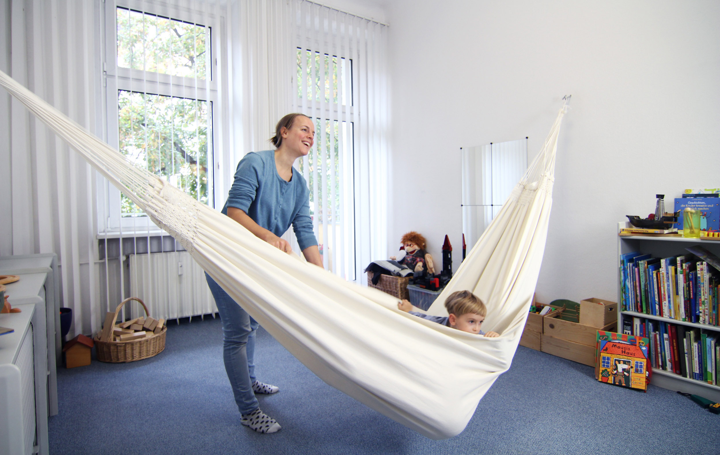Lachende Logopädin schaukelt einen Jungen in der Hängematte: Therapieraum für Kinder mit Bücherregal und Spielsachen in der Praxis in Berlin-Spandau